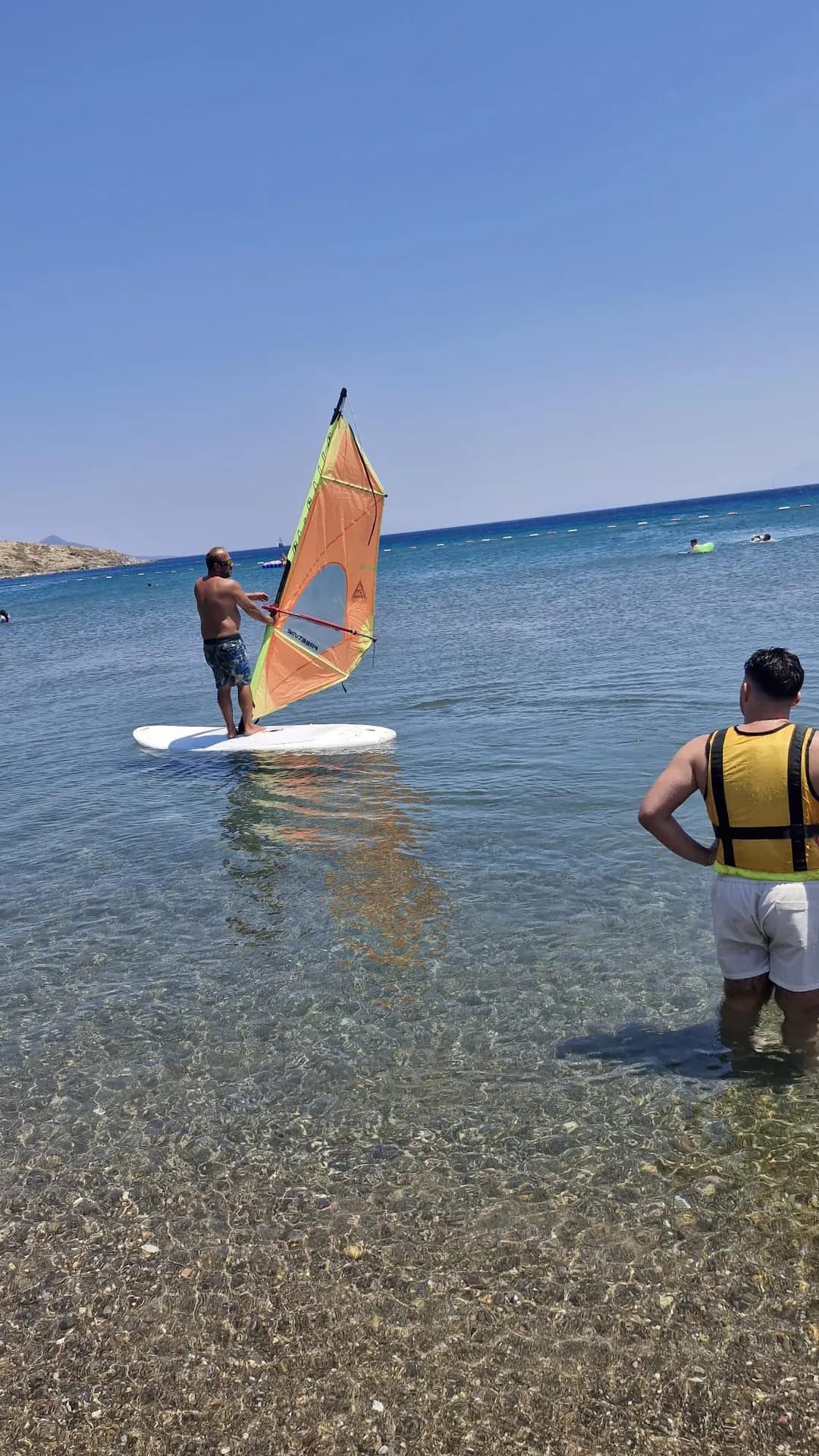 Sığ suda turuncu yelkenle dengede duran windsurf katılımcısı